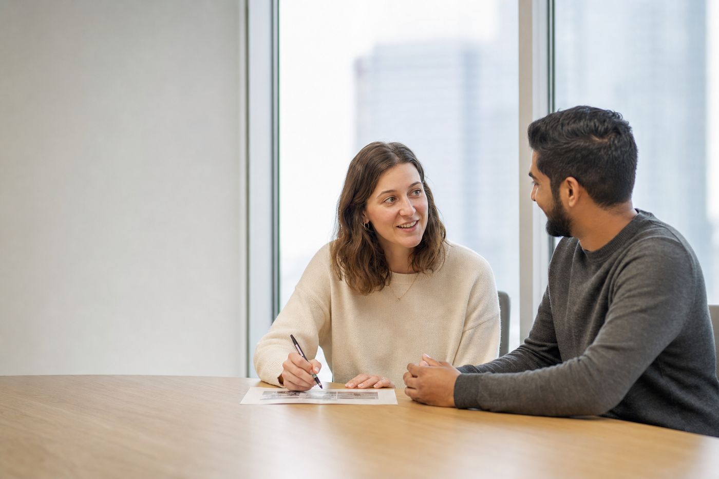 Two people at a table in a bright office, mid conversation over a printed page.